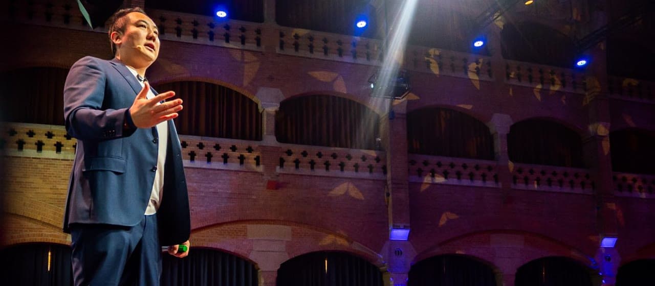 John Lee on stage in a brick concert hall, mid-talk